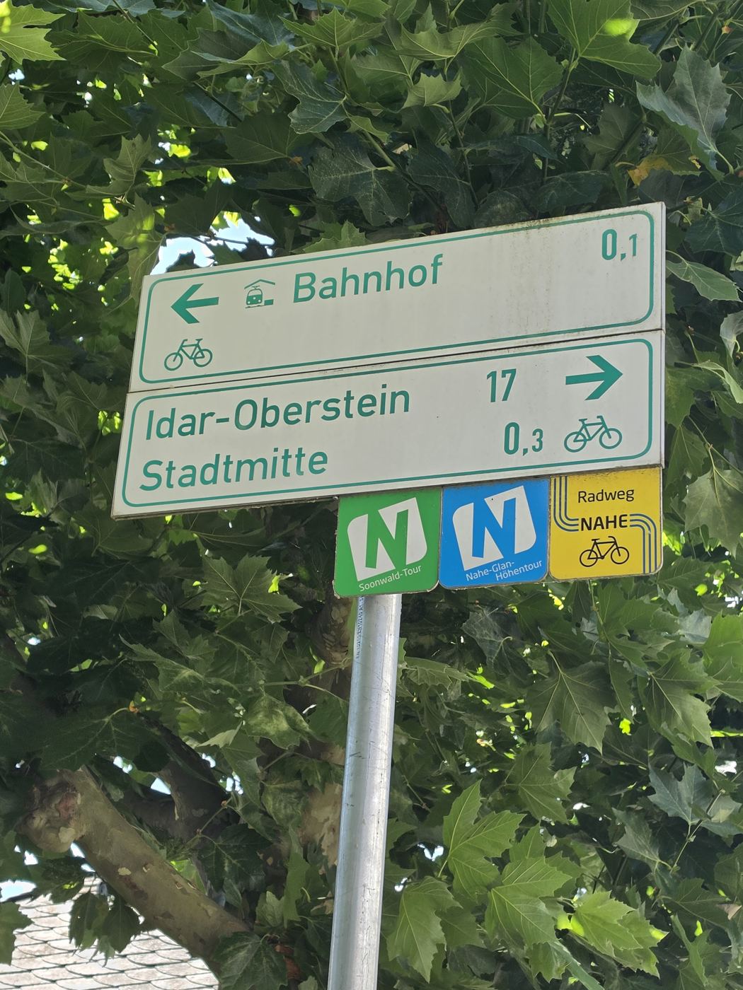 Radwegweiser am Bahnhof Kirn mit Richtungen Bahnhof, Stadtmitte und Idar-Oberstein, dahinter Platanenlaub
